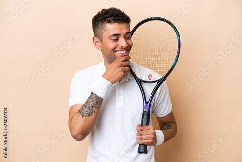 Young brazilian handsome man playing tennis isolated on beige background looking to the side and smiling © luismolinero