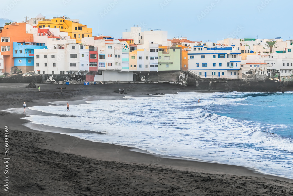Waves wash black sand with white foam at Playa Maria Jimenez beach in Puerto de la Cruz during a storm, Spain. Several people rest in Atlantic Ocean on the backdrop of beautiful buildings in Canaries