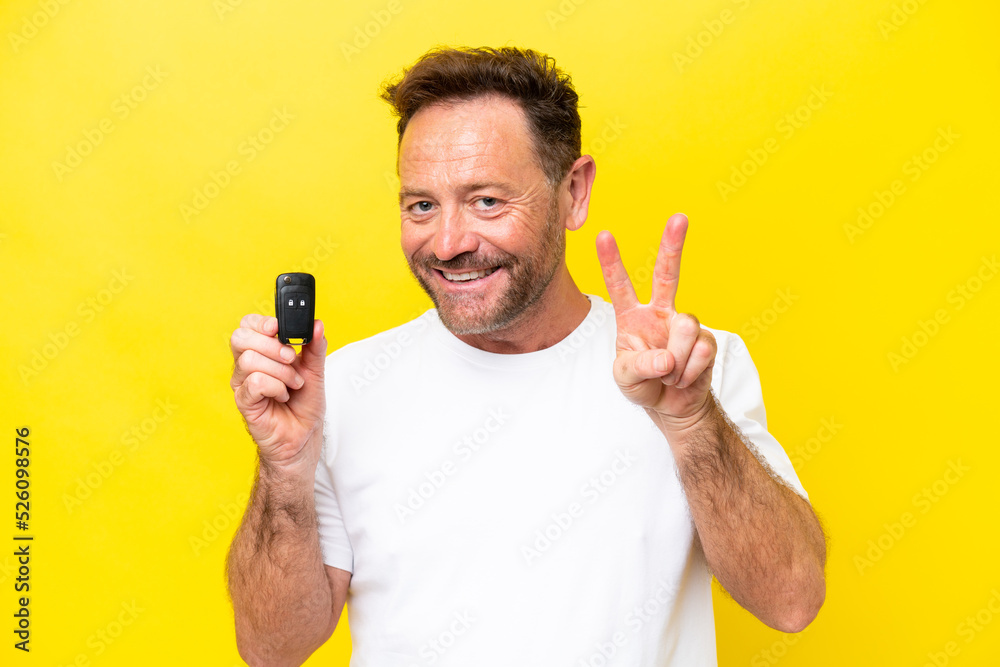 Middle age caucasian man holding car keys isolated on yellow background smiling and showing victory sign Middle age caucasian man holding car keys isolated on yellow background smiling and showing victory sign