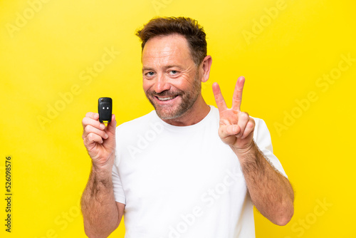 Middle age caucasian man holding car keys isolated on yellow background smiling and showing victory sign