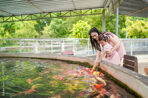 Asian young woman and her daughter holding together with a milk bottle feeding a school of Japanese carp fish, Koi fish in a pond.