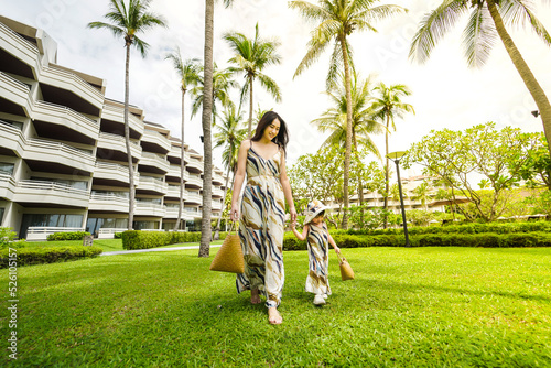 Asian young woman and her daughter in a fashion clothes with weave handbag enjoying walking along a grass field on vacation.