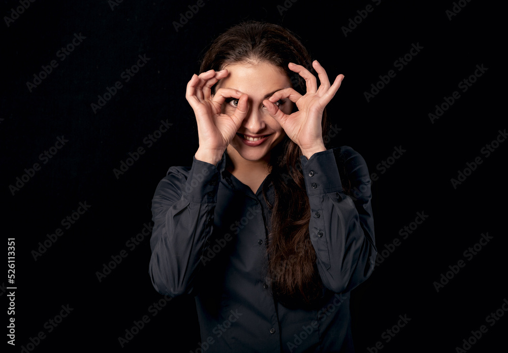 Beautiful funny business woman showing the watching binoculars and peep sign holding hands near the eyes with big eyes on black background. Binoculars fingers gesture. Beautiful funny business woman showing the watching binoculars and peep sign holding hands near the eyes with big eyes on black background. Binoculars fingers gesture.