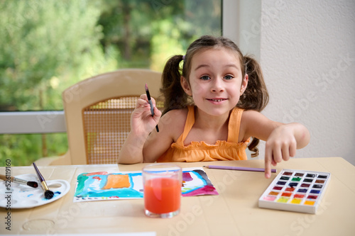 Happy little girl sitting at table and cutely smiling at camera while painting with watercolors and paintbrush. Kids education and development. Drawing. Art classes and courses for telented children photo