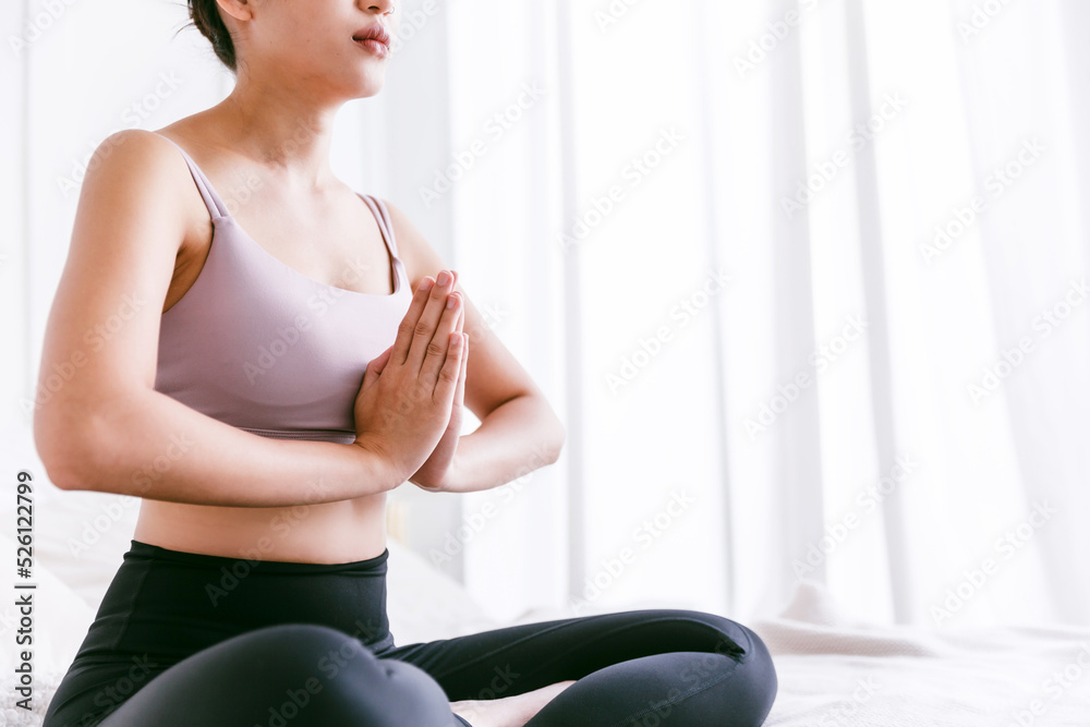Crop image of young attractive sporty Asian woman practicing yoga on the bed, doing Ardha Padmasana exercise, meditating in Half Lotus pose with namaste, indoor working out at home. Crop image of young attractive sporty Asian woman practicing yoga on the bed, doing Ardha Padmasana exercise, meditating in Half Lotus pose with namaste, indoor working out at home.