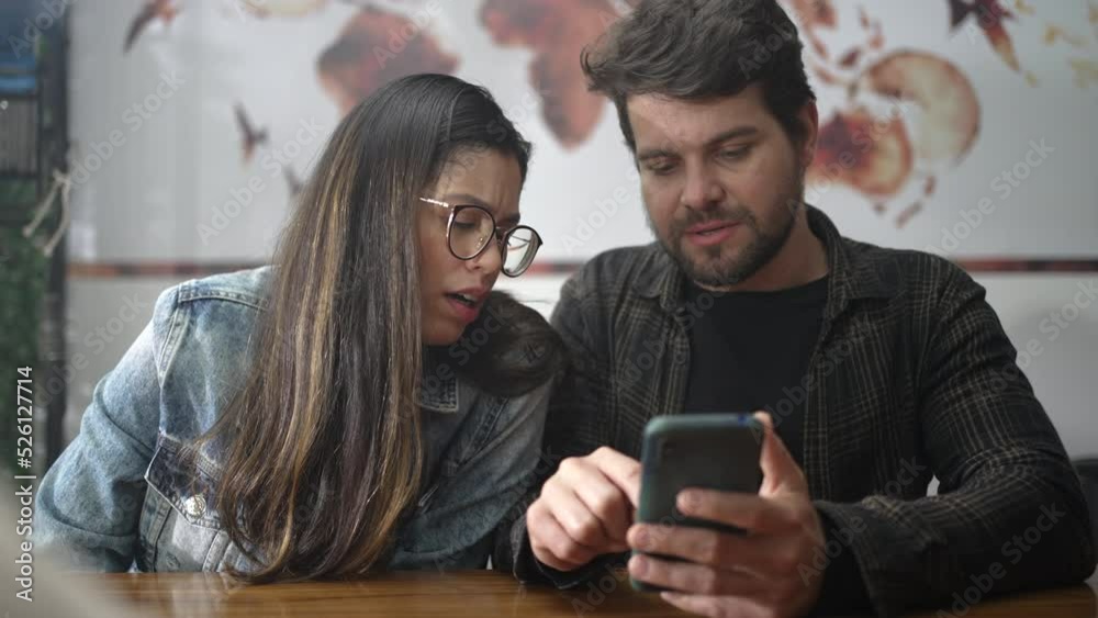 Couple looking at smartphone screen seated at coffee shop. People laughing and smiling over video content online. Young man showing phone screen to girl Couple looking at smartphone screen seated at coffee shop. People laughing and smiling over video content online. Young man showing phone screen to girl