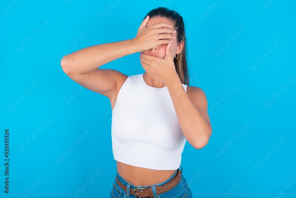 beautiful brunette woman wearing white tank top over blue background Covering eyes and mouth with hands, surprised and shocked. Hiding emotions.