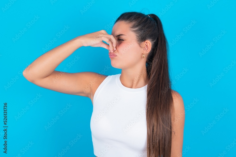 beautiful brunette woman wearing white tank top over blue background smelling something stinky and disgusting, intolerable smell, holding breath with fingers on nose. Bad smell - obrazy, fototapety, plakaty 