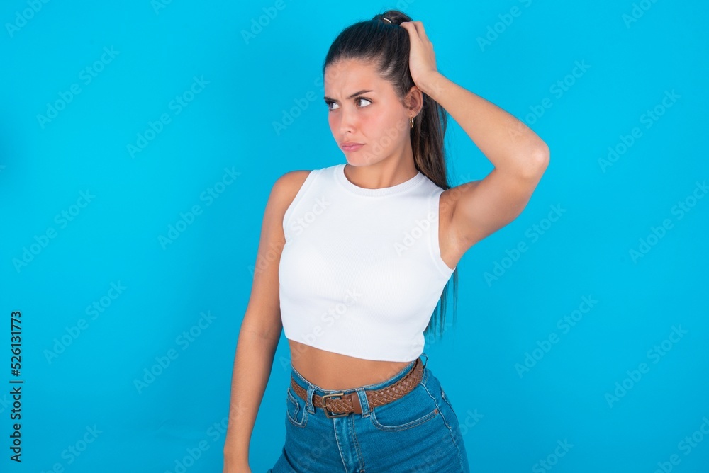 beautiful brunette woman wearing white tank top over blue background confuse and wonder about question. Uncertain with doubt, thinking with hand on head. Pensive concept. beautiful brunette woman wearing white tank top over blue background confuse and wonder about question. Uncertain with doubt, thinking with hand on head. Pensive concept.