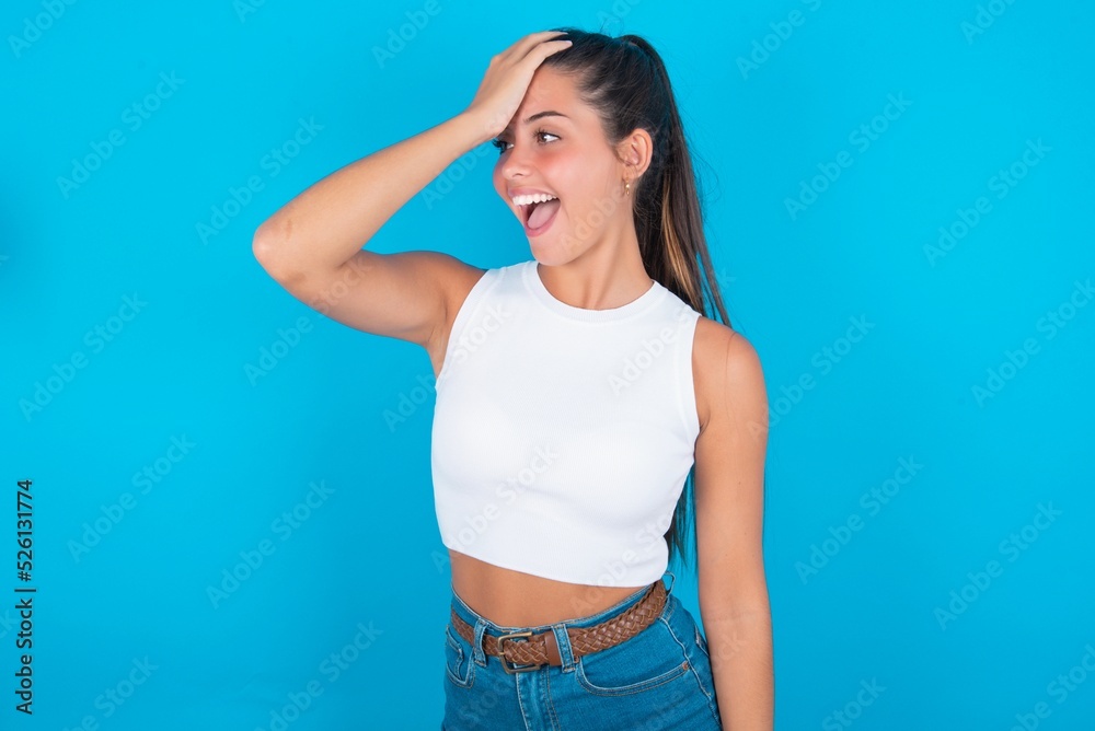 beautiful brunette woman wearing white tank top over blue background surprised with hand on head for mistake, remember error. Forgot, bad memory concept. beautiful brunette woman wearing white tank top over blue background surprised with hand on head for mistake, remember error. Forgot, bad memory concept.