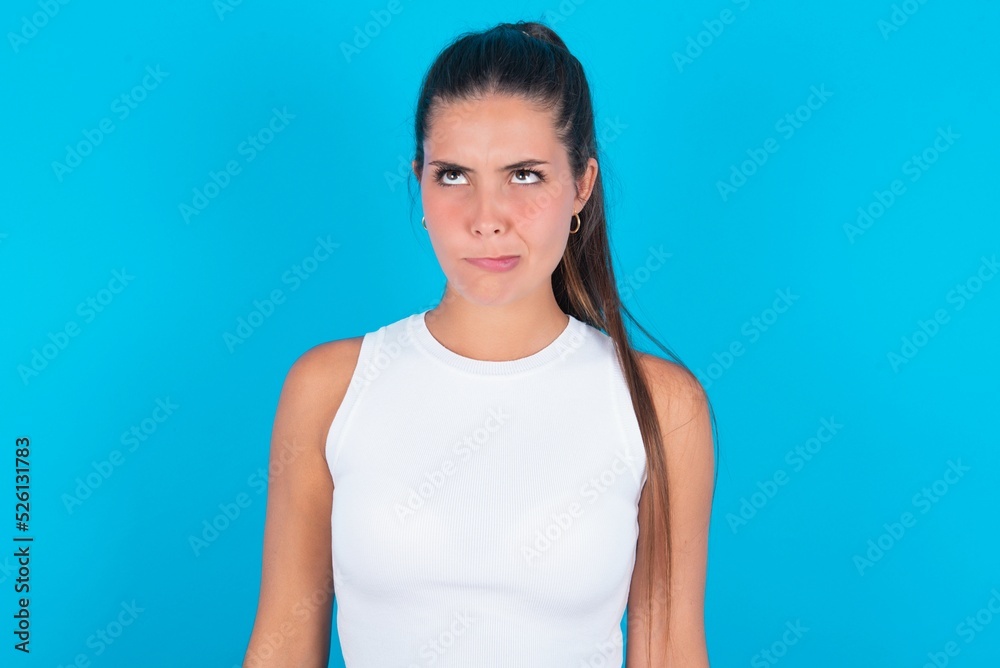 beautiful brunette woman wearing white tank top over blue background making grimace and crazy face, screaming out of control, funny lunatic expressing freedom and wild. beautiful brunette woman wearing white tank top over blue background making grimace and crazy face, screaming out of control, funny lunatic expressing freedom and wild.
