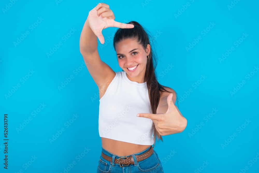 beautiful brunette woman wearing white tank top over blue background making finger frame with hands. Creativity and photography concept. beautiful brunette woman wearing white tank top over blue background making finger frame with hands. Creativity and photography concept.