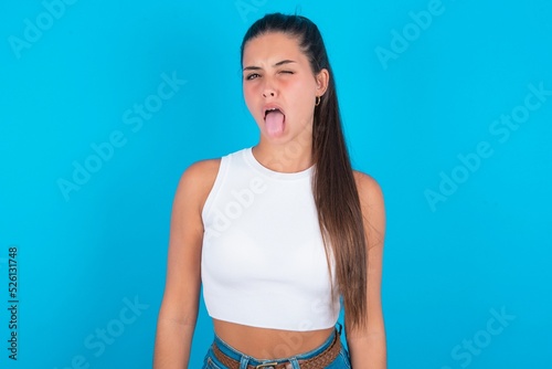 beautiful brunette woman wearing white tank top over blue background sticking tongue out happy with funny expression. Emotion concept. © Roquillo