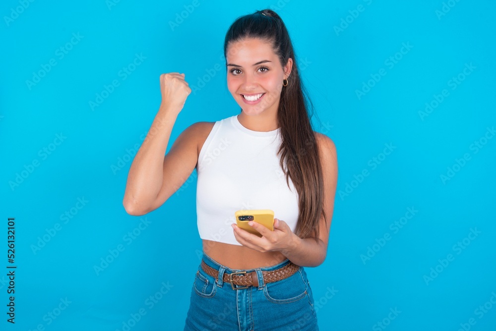Portrait of beautiful brunette woman wearing white tank top over blue background holding in hands cell celebrating Portrait of beautiful brunette woman wearing white tank top over blue background holding in hands cell celebrating