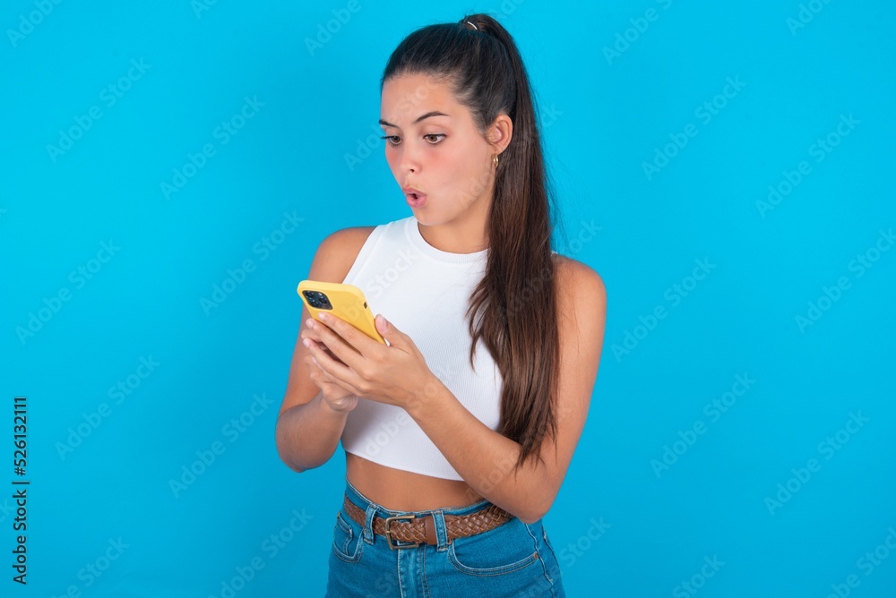 Focused beautiful brunette woman wearing white tank top over blue background use smartphone reading social media news, or important e-mail Focused beautiful brunette woman wearing white tank top over blue background use smartphone reading social media news, or important e-mail