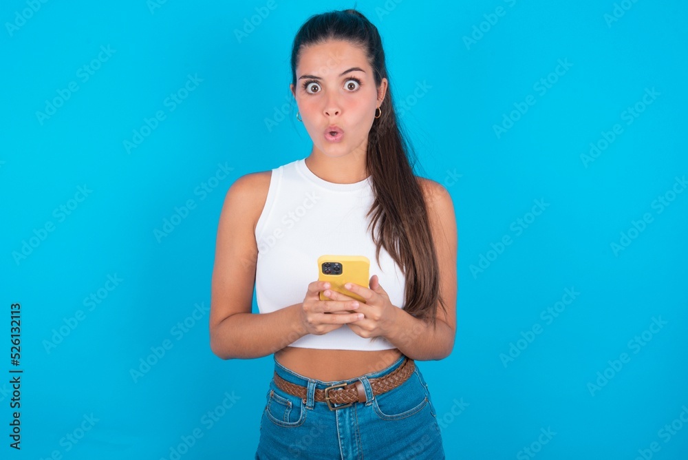 Shocked beautiful brunette woman wearing white tank top over blue background opens mouth hold phone reading advert unbelievable big shopping prices Shocked beautiful brunette woman wearing white tank top over blue background opens mouth hold phone reading advert unbelievable big shopping prices