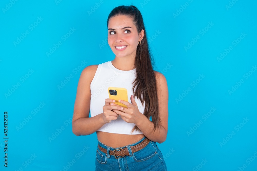 beautiful brunette woman wearing white tank top over blue background hold telephone hands read good youth news look empty space advert