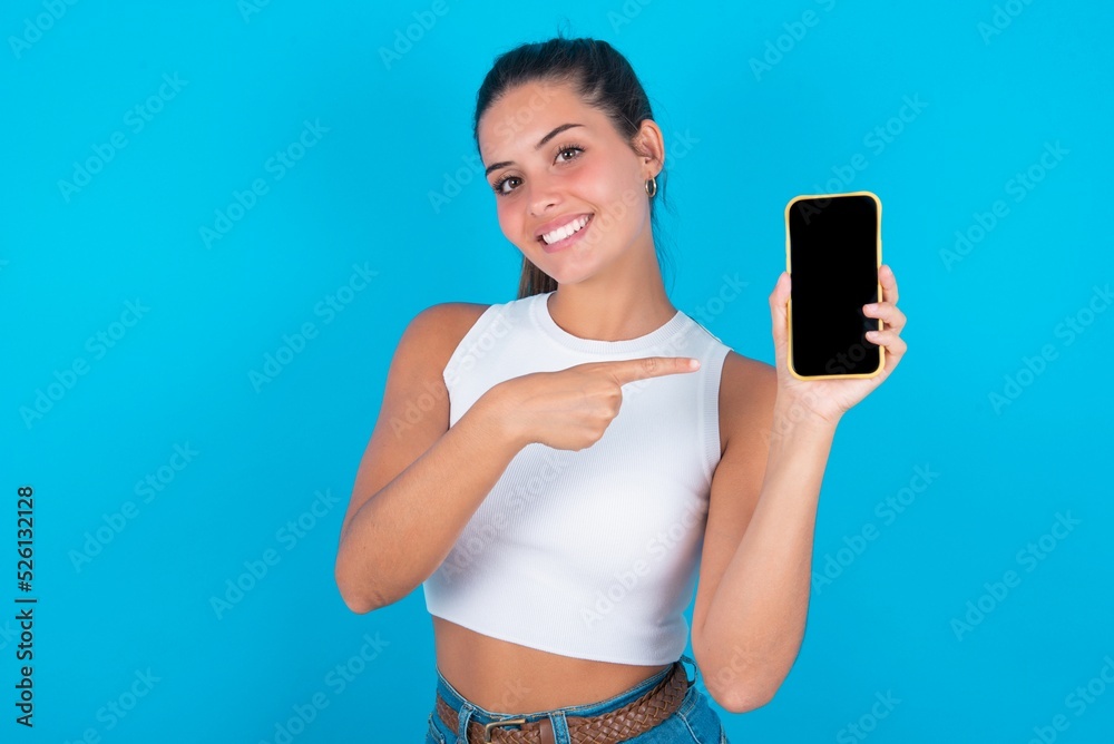 Smiling beautiful brunette woman wearing white tank top over blue background Mock up copy space. Pointing index finger on mobile phone with blank empty screen