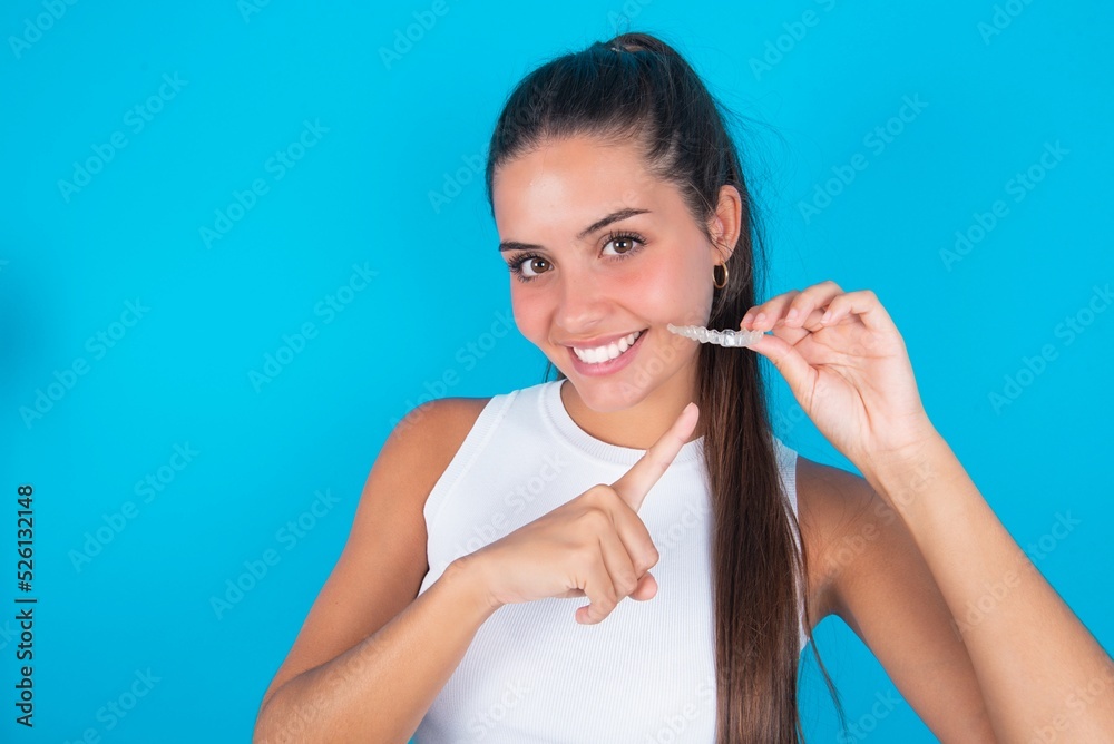 beautiful brunette woman wearing white tank top over blue background holding an invisible aligner and pointing at it. Dental healthcare and confidence concept. beautiful brunette woman wearing white tank top over blue background holding an invisible aligner and pointing at it. Dental healthcare and confidence concept.