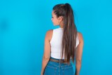 The back side view of a beautiful brunette woman wearing white tank top over blue background. Studio Shoot.
