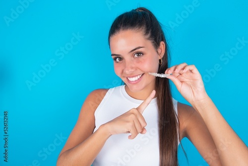 beautiful brunette woman wearing white tank top over blue background holding an invisible aligner and pointing at it. Dental healthcare and confidence concept.