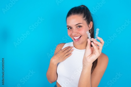 Happy beautiful brunette woman wearing white tank top over blue background holding and showing at camera an invisible aligner while laughing. Dental healthcare and confidence concept. photo