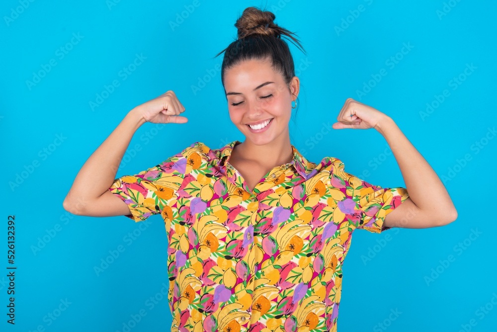Strong powerful beautiful brunette woman wearing colourful shirt over blue background toothy smile, raises arms and shows biceps. Look at my muscles! Strong powerful beautiful brunette woman wearing colourful shirt over blue background toothy smile, raises arms and shows biceps. Look at my muscles!