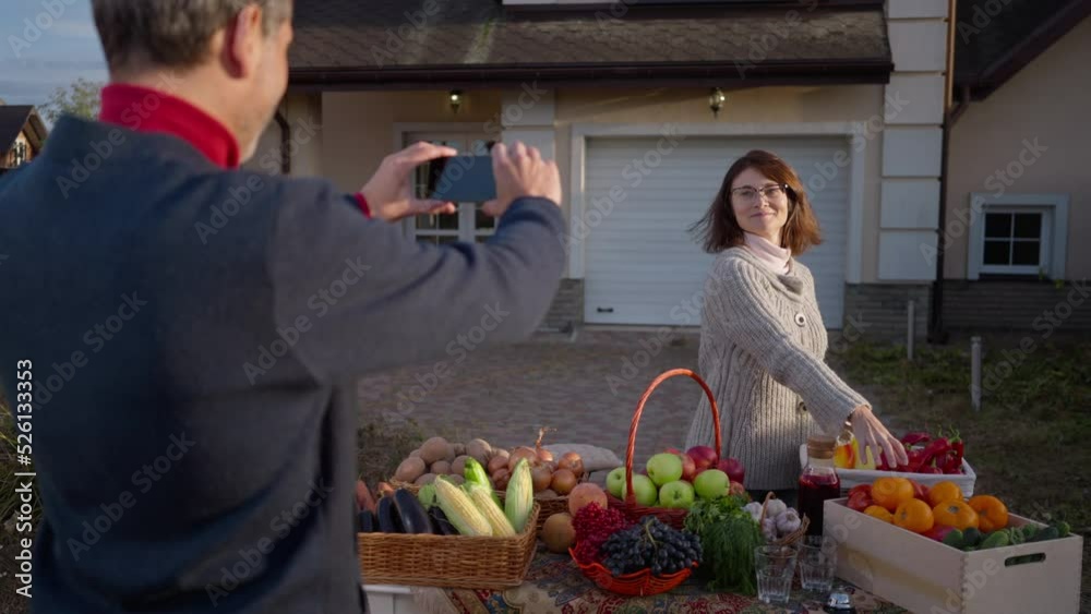 Back view man taking photo of woman posing with organic local products on farm market. Shooting over shoulder of loving husband supporting wife selling healthful fruits and vegetables Back view man taking photo of woman posing with organic local products on farm market. Shooting over shoulder of loving husband supporting wife selling healthful fruits and vegetables