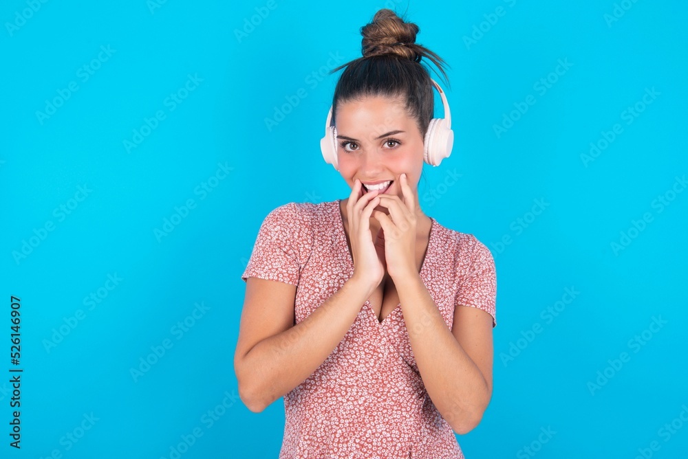 Shocked beautiful brunette woman wearing floral dress over blue background stares fearful at camera keeps mouth widely opened wears wireless stereo headphones on ears Shocked beautiful brunette woman wearing floral dress over blue background stares fearful at camera keeps mouth widely opened wears wireless stereo headphones on ears
