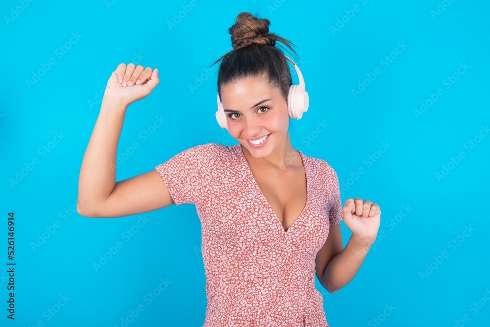 Fototapeta premium Carefree beautiful brunette woman wearing floral dress over blue background with toothy smile raises arms dances carefree moves with rhythm of music listens music from playlist via headphones