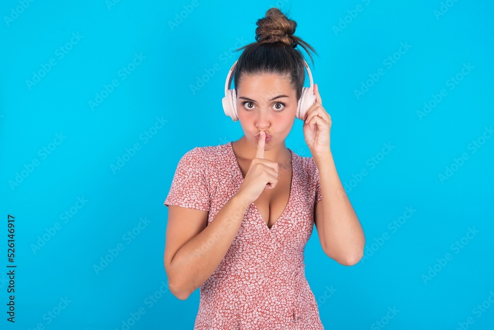 beautiful brunette woman wearing floral dress over blue background making hush gesture with finger on her lips wearing wireless headphones. Be quiet. beautiful brunette woman wearing floral dress over blue background making hush gesture with finger on her lips wearing wireless headphones. Be quiet.