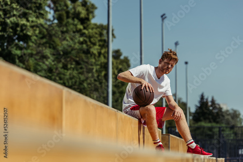 young basketball player with ball sitting on stadium on blurred foreground