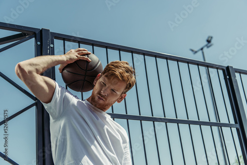low angle view of redhead man in white t-shirt standing with ball near fence photo