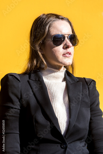 Executive entrepreneur woman, wearing black jacket and wool sweater and sunglasses, on yellow background in the street, with copy space. Entrepreneur woman concept.