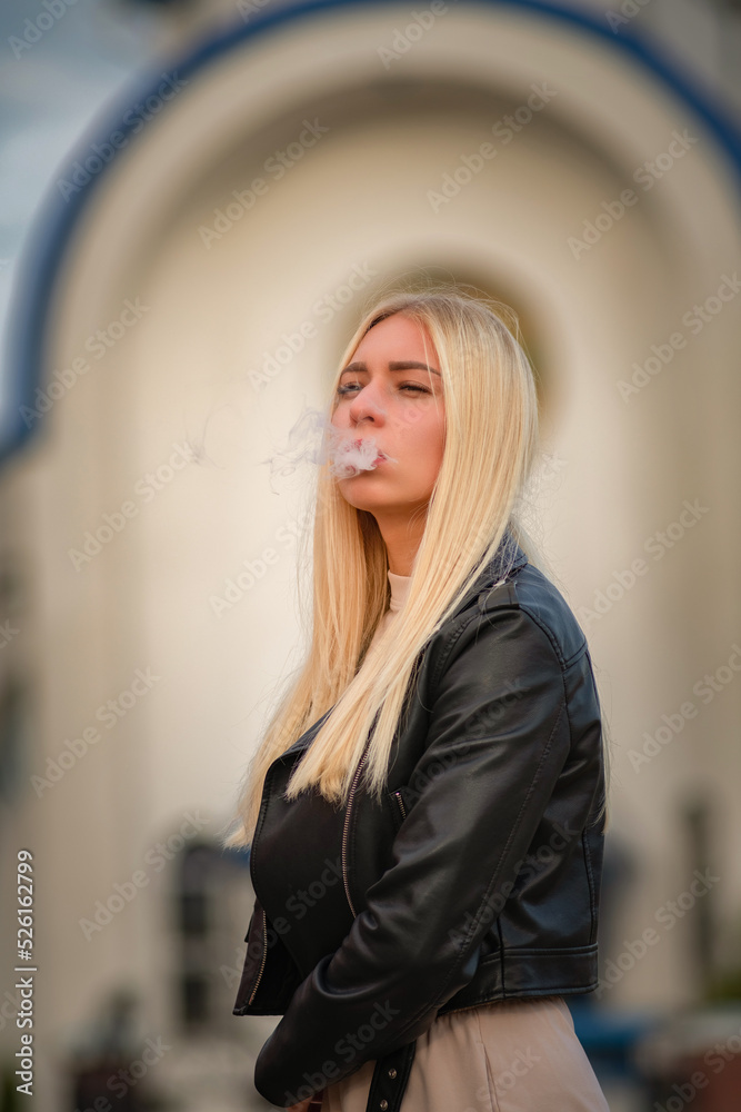Portrait of a young beautiful fair-haired girl letting out cigarette smoke from her mouth.
