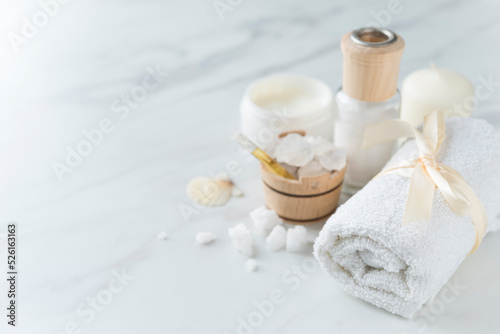 Spa composition with towel, small wooden bowl with coarse sea salt, vial of oil, candle, skin cream, sea shell on the light marble background. Copy space. Top view