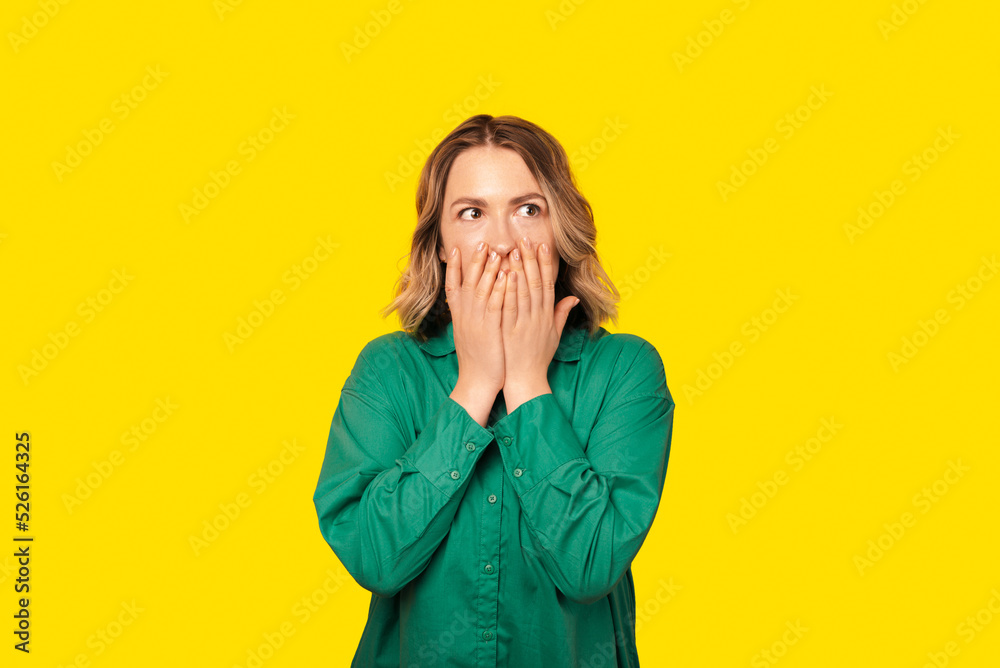 Shocked woman is looking away with her hands covering her mouth, wearing a green shirt over yellow wall Shocked woman is looking away with her hands covering her mouth, wearing a green shirt over yellow wall