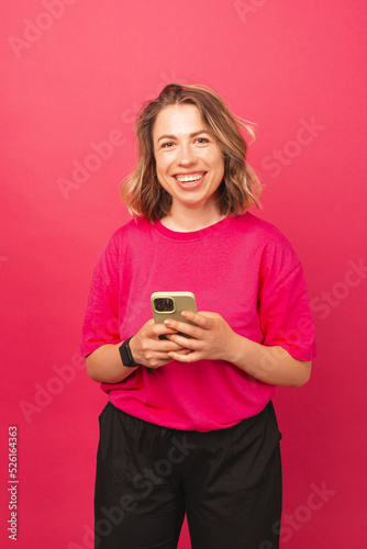Smiling woman is holding her phone texting someone wearing a pink shirt over a wall with the same color