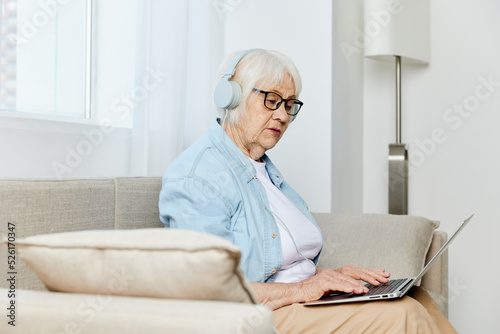 a pleasant, happy elderly woman is sitting on a beige sofa and holding a laptop on her lap, communicating via video using headphones © Tatiana