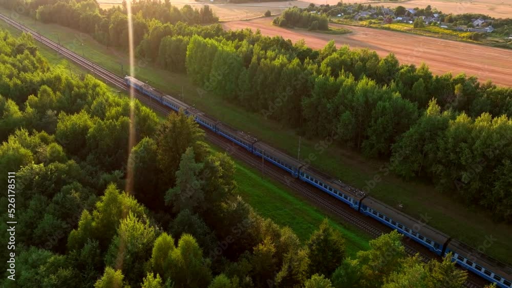 Passenger train on railway from forest, aerial view. Train with passenger cars rides along forests on sunset. Drone view of an electric passenger locomotive with wagons in moving on railroad.