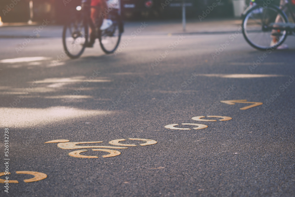 Obraz premium crowded inner city junction with modern bicycle route symbols painted in orange on asphalt. selective focus on single symbol with blurred bicycle riders in background. urban mobility concept