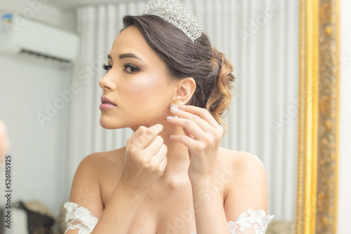 Jovem mulher vestida de noiva, de frente ao espelho, colocando seus brincos. photo