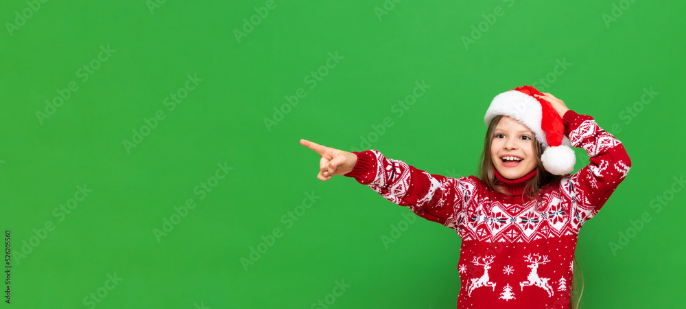 A little girl in a Santa Claus hat and a festive sweater points to your advertisement and smiles broadly on a green isolated background. Copy space.