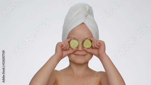 Beautiful young smiling child girl holding slices of cucumber, covering eyes. Teenager kid face skin care treatment, natural cosmetics. Female portrait on white background. Perfect fresh clean skin