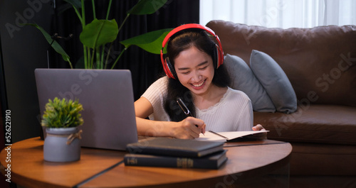 Smiling Asian young female in headphone making notes on notebook, learning online courses on laptop. E-learning education concept.