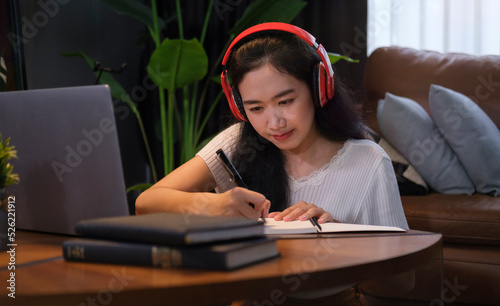 Pretty young woman in headphones writing important information on notebook during online lecture via laptop.