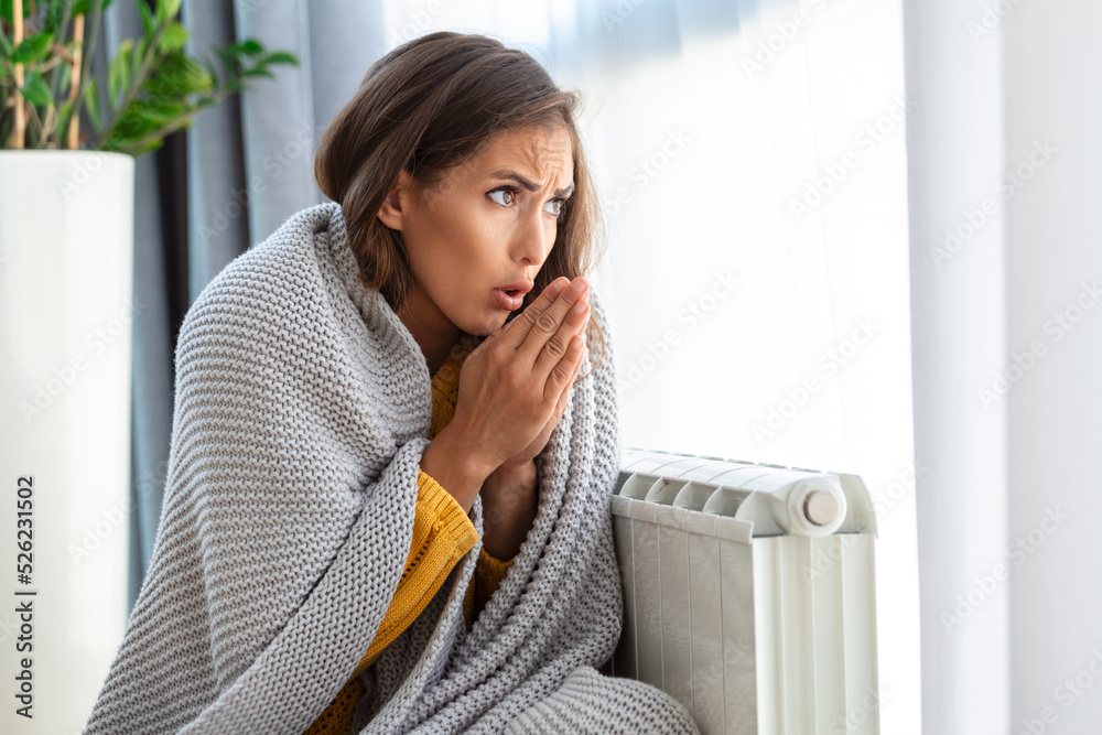Unwell woman renter in blanket sit in cold living room hand on old radiator.suffer  from lack of heat . Unhealthy young woman struggle from chill freeze at  home. No heating concept. Stock