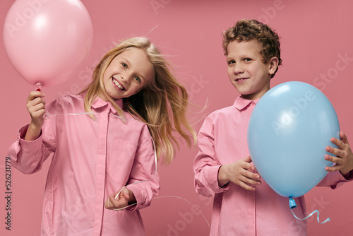 two joyful, cute children stand on a pink background holding pink and blue balloons in their hands, smiling joyfully at the camera. Horizontal studio photography with empty space
