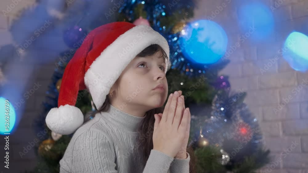 Alone girl pray at Christmas night. An alone girl in Santa hat stay in praying position by the Christmas tree in the room. Alone girl pray at Christmas night. An alone girl in Santa hat stay in praying position by the Christmas tree in the room.