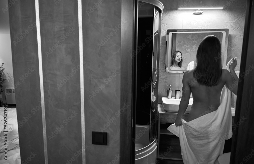 Young naked woman in towel, looking in mirror in the bathroom. Black and white image. Young naked woman in towel, looking in mirror in the bathroom. Black and white image.